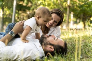 Familia de tres jugando y riendo en el césped con luz natural, el padre acostado. Fotografo de familias en Vigo, momentos no forzados.