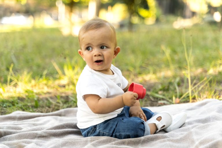 Bebé sentado solo sobre una manta en el césped, mirando a la cámara, con luz natural. Fotografo de bebes en Vigo, gesto natural.