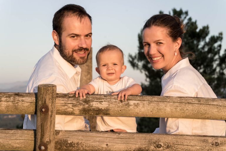 Familia de tres posando junto a una valla de madera al atardecer, con luz natural. Fotografo de familias en Galicia, retrato relajado.