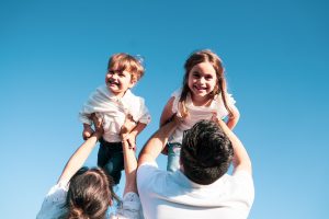 Padres levantando a sus dos hijos en el aire, jugando con fondo de cielo azul. Fotografo infantil en Galicia, momentos