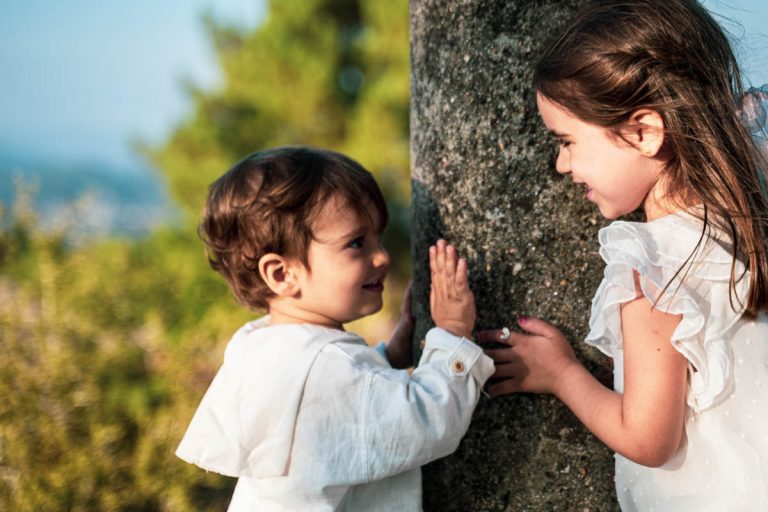 Dos hermanos pequeños interactuando junto a un árbol, el sol creando un brillo. Fotografo de familias en Vigo, Galicia.